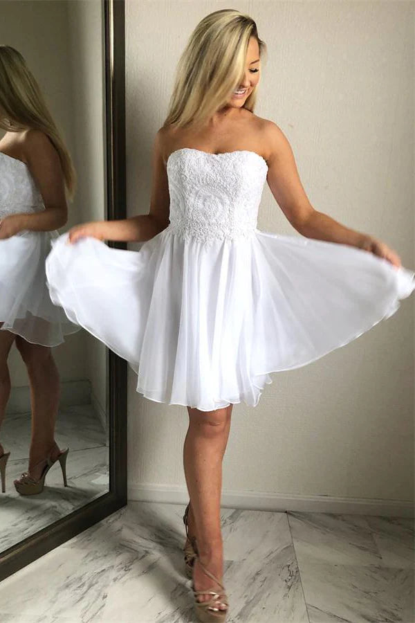 A woman in a white chiffon homecoming dress with sweetheart lace detailing, posing in a room with a mirror.