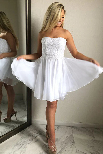 A woman in a white chiffon homecoming dress with sweetheart lace detailing, posing in a room with a mirror.